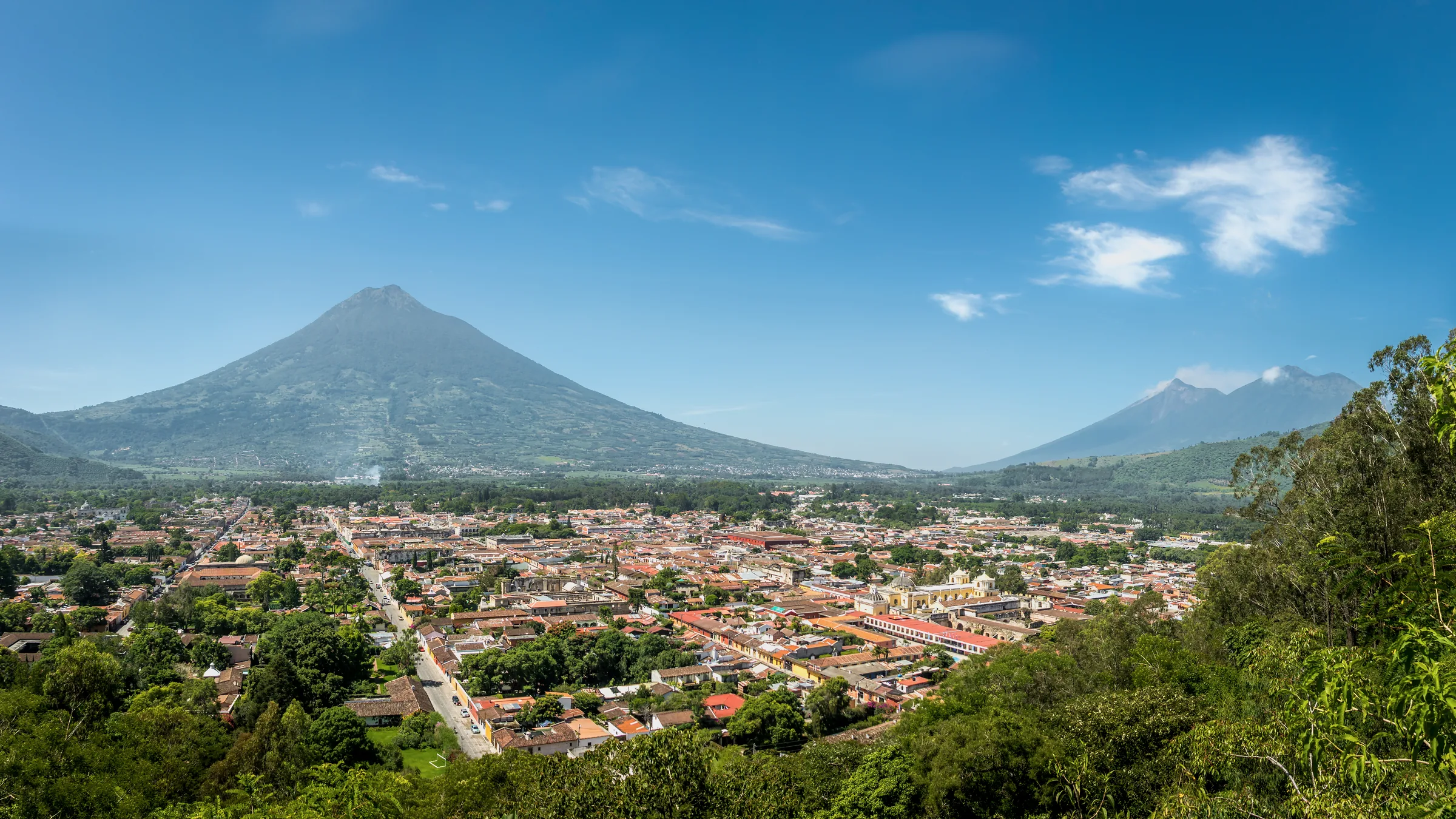 Tejados de Antigua Guatemala al pie de un volcán, vistos desde una ladera con vegetación.